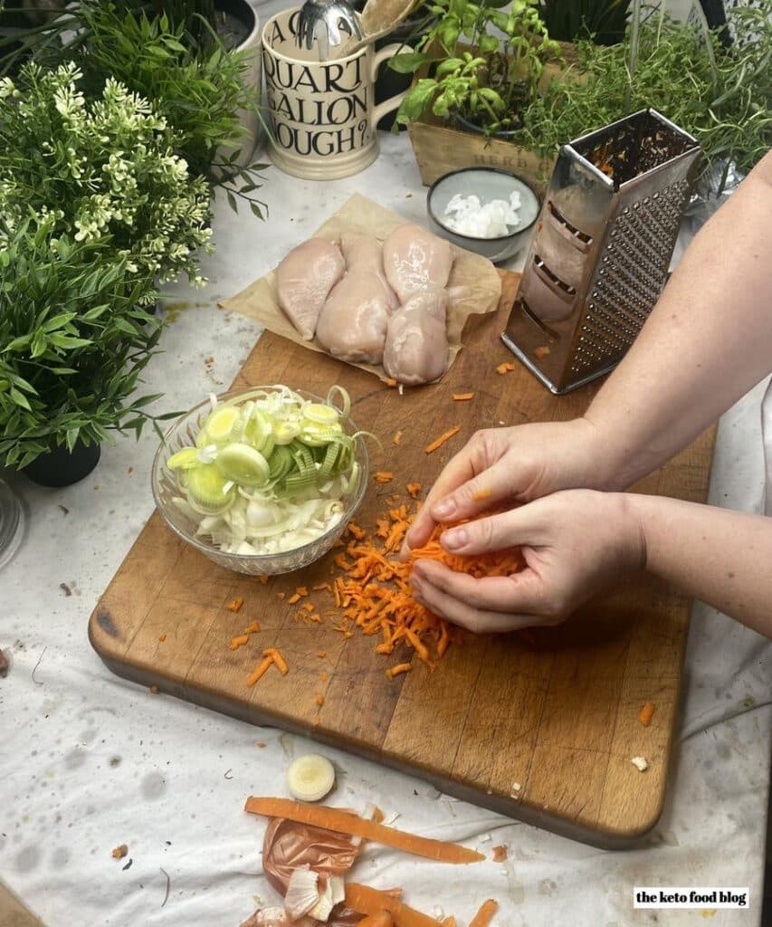 Grated carrot on a wooden chopping board being scooped into a bowl of vegetables. 