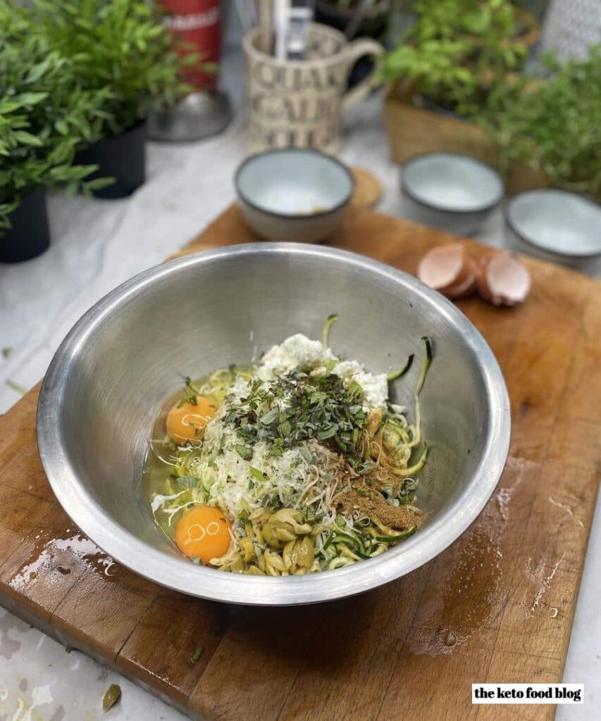 A metal mixing bowl full of eggs, zucchini and herbs to make fritters