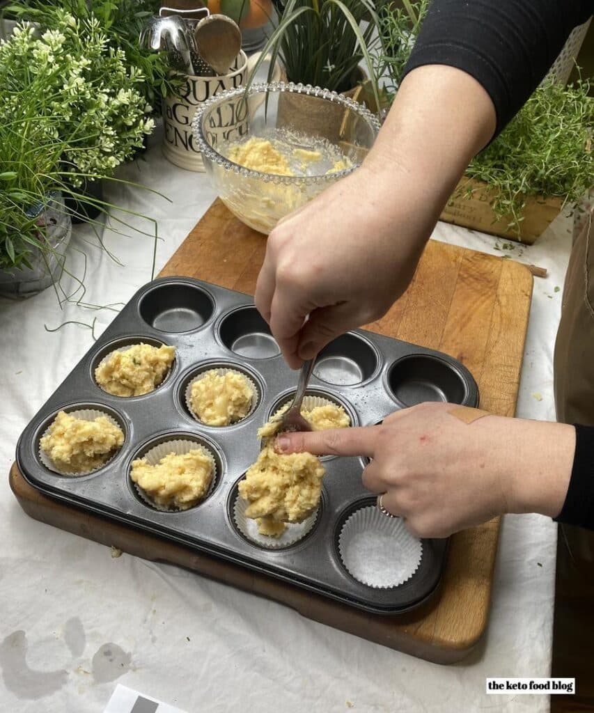 Spooning savoury muffin mixture into muffin cases in a muffin tin on a wooden chopping board. 