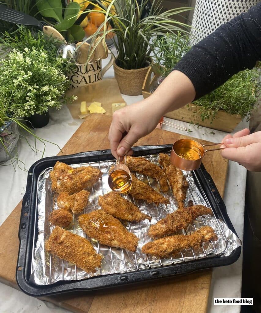 Drizzling olive oil from a copper spoon over breaded and seasoned chicken strips on a baking tray